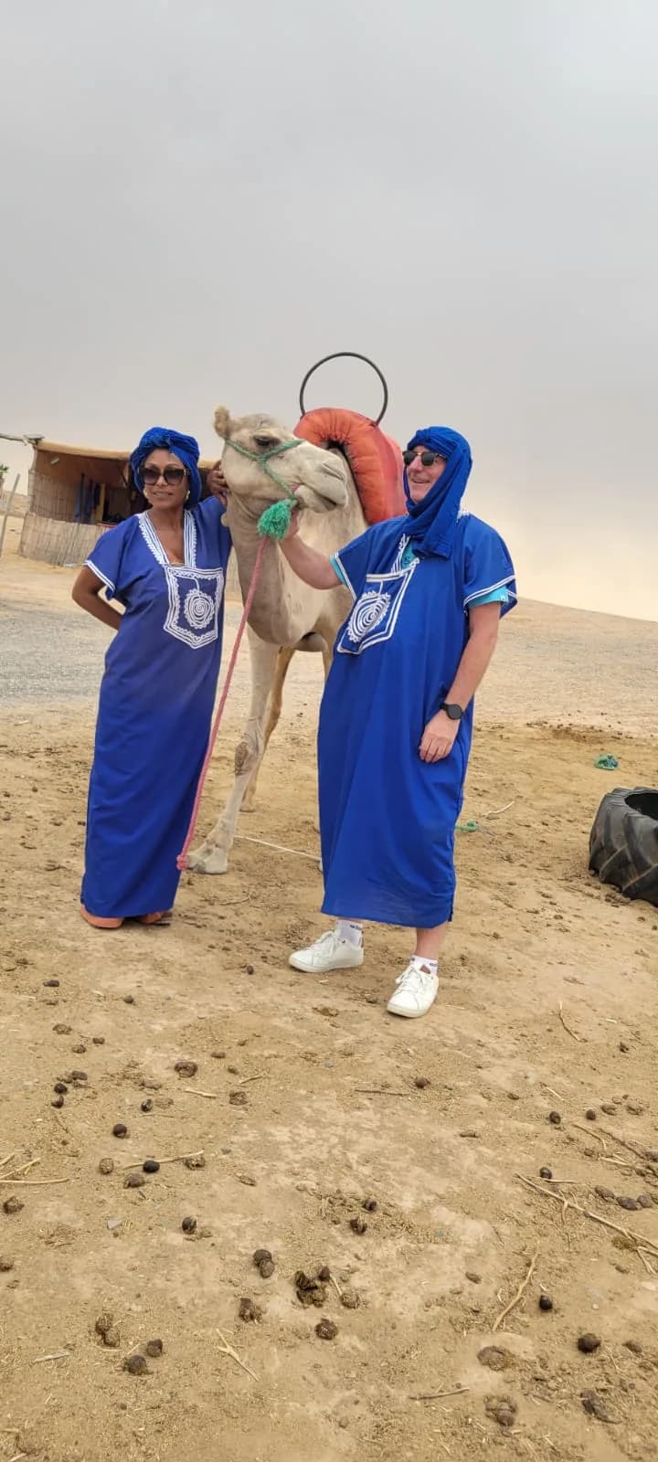 Two guests in traditional blue robes standing beside a camel in the Agafay Desert