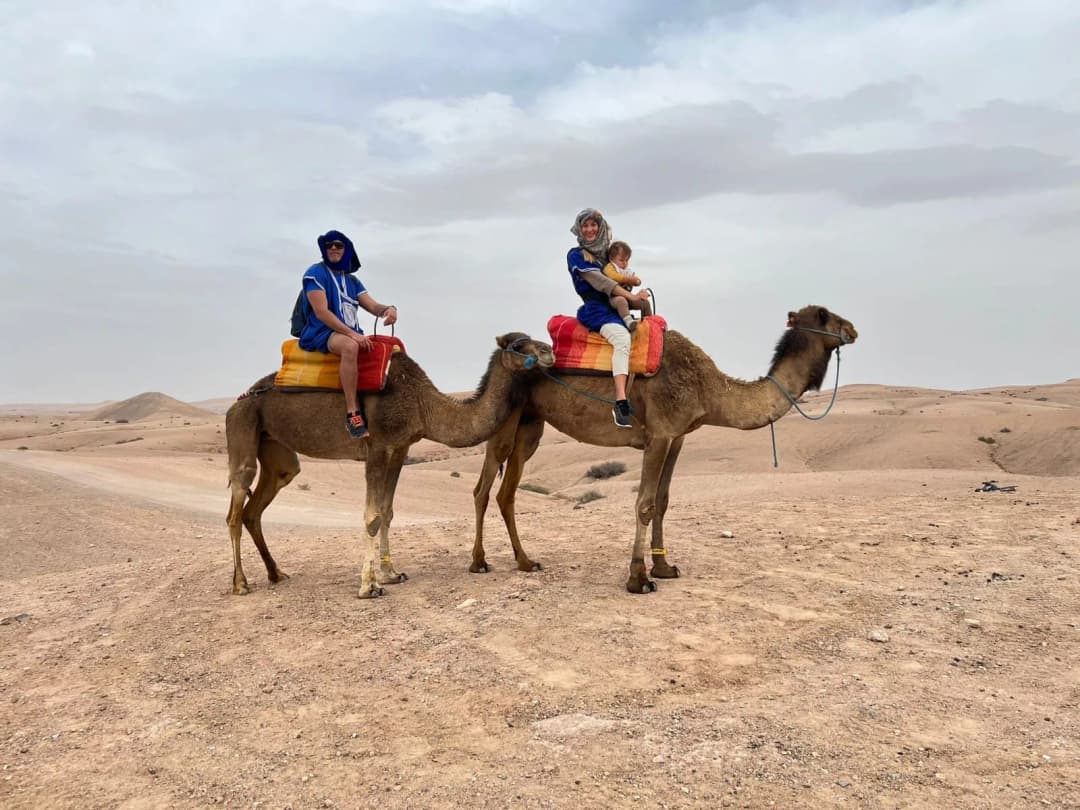Two riders, an adult and a child, on camels crossing pale stony desert under a cloudy sky