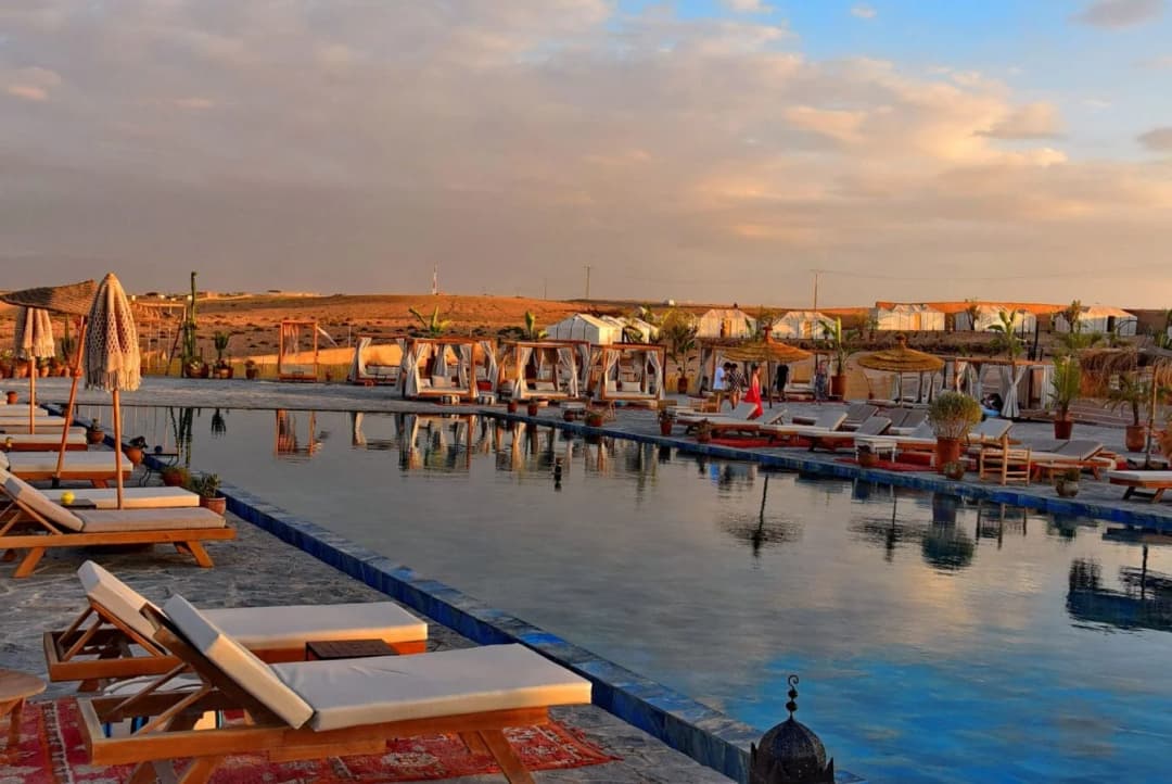 An outdoor pool at dusk with sun loungers, curtained cabanas and white tents behind it in the Agafay Desert